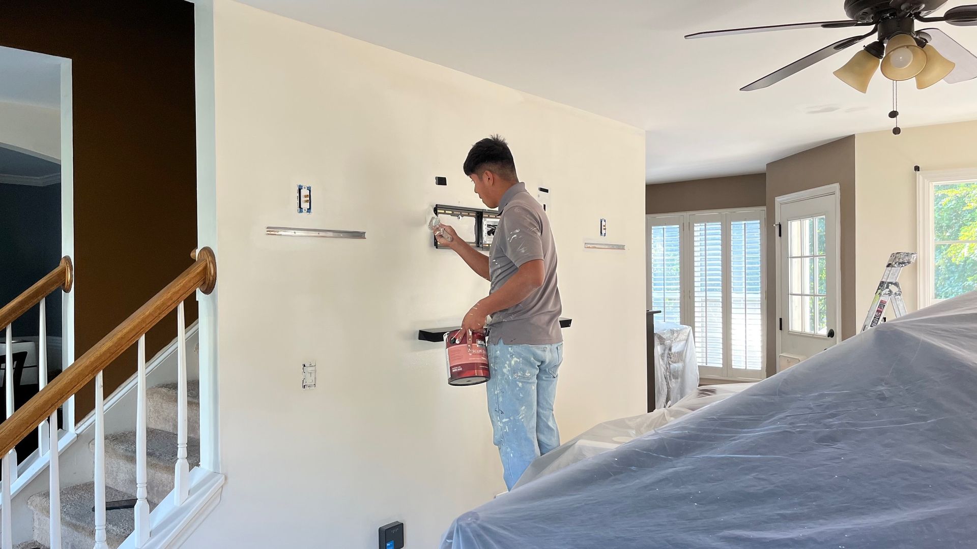 Person painting a wall in a home. They are standing on covered furniture. The wall is off-white and brown.