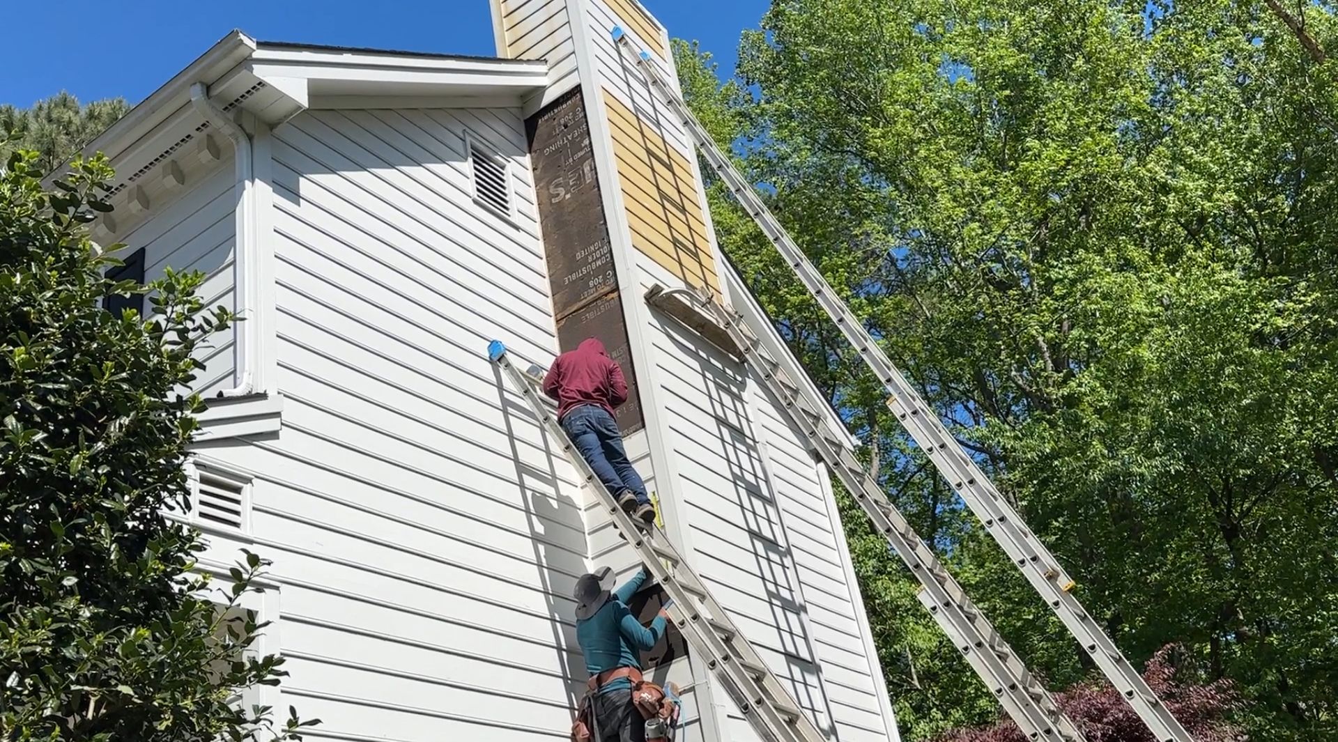Two workers on ladders repair a chimney on a white house; one removes siding.