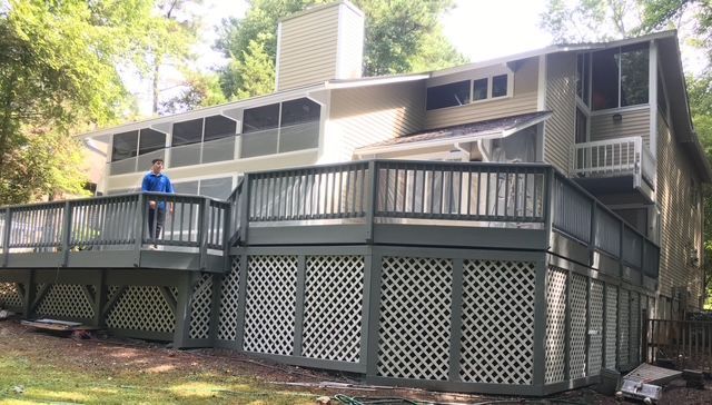 Two-story house with green deck, lattice skirting, person standing on the deck, and a wooded background.