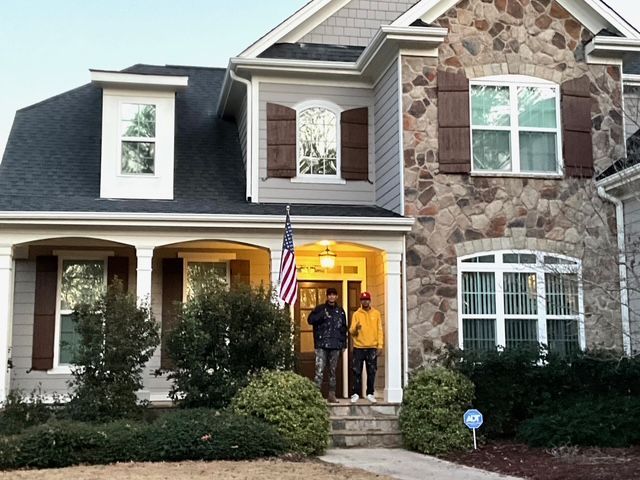 Two people stand on the porch of a large house with stone and siding. An American flag hangs.