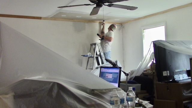 Person on a ladder installing trim on a white ceiling, room is covered in plastic, TV and computer visible.