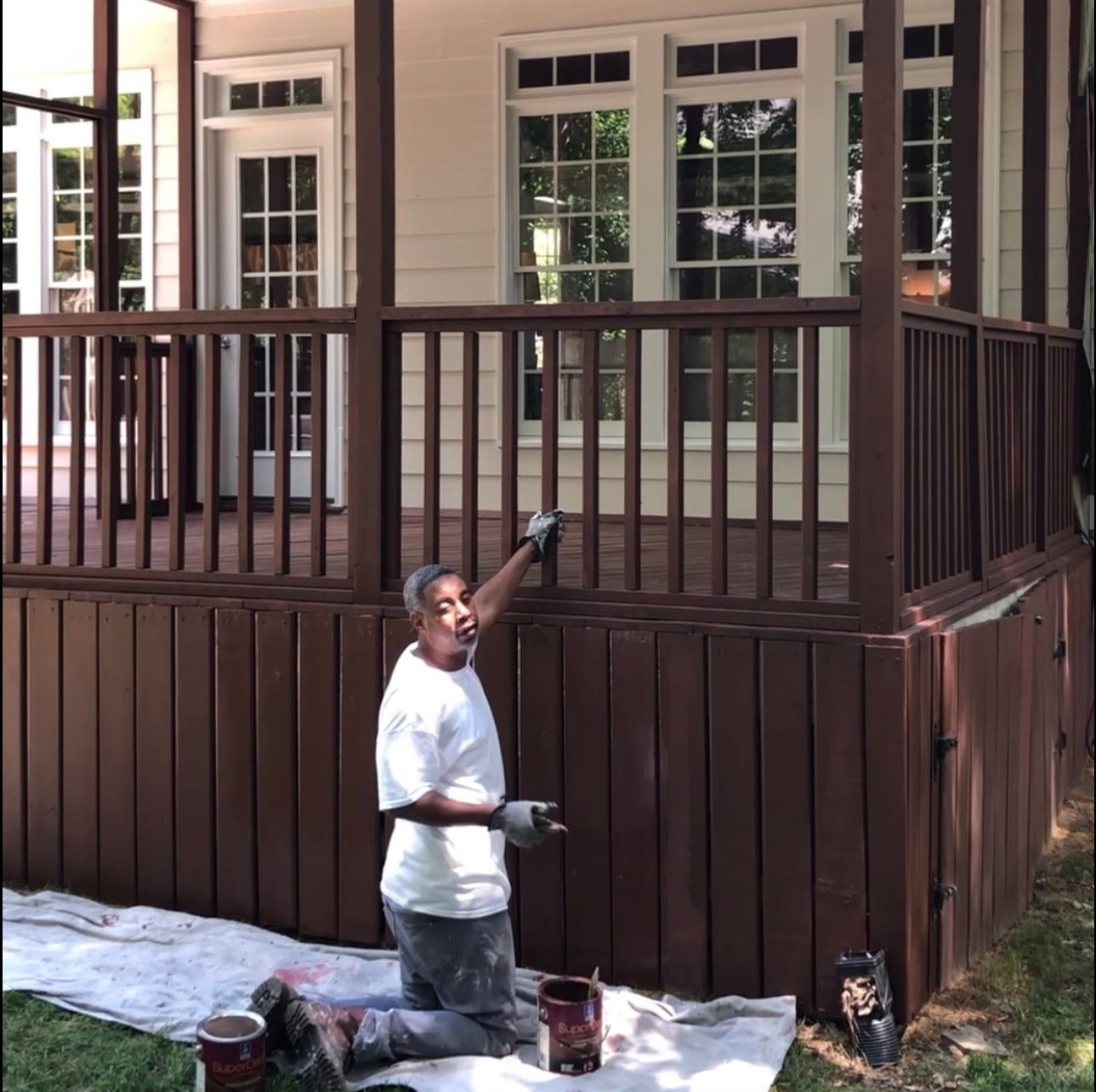 Man paints a brown deck railing and side paneling while kneeling outdoors.