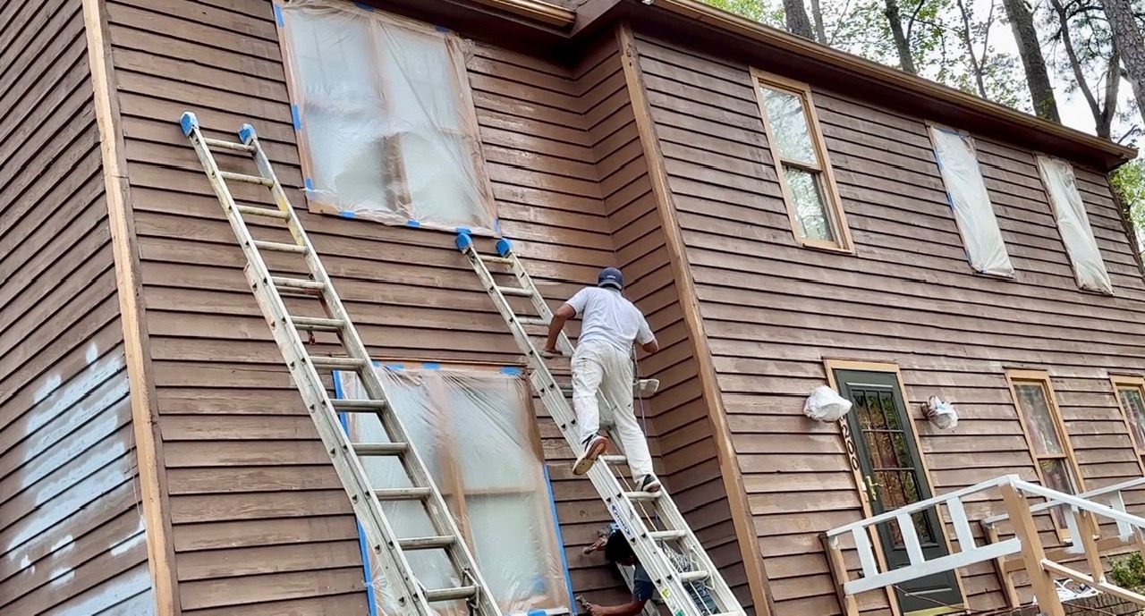 Person painting brown house siding from ladders. Windows covered with plastic.