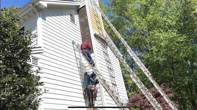 Two people on ladders next to a tall white house, one near the roof, working on the side.
