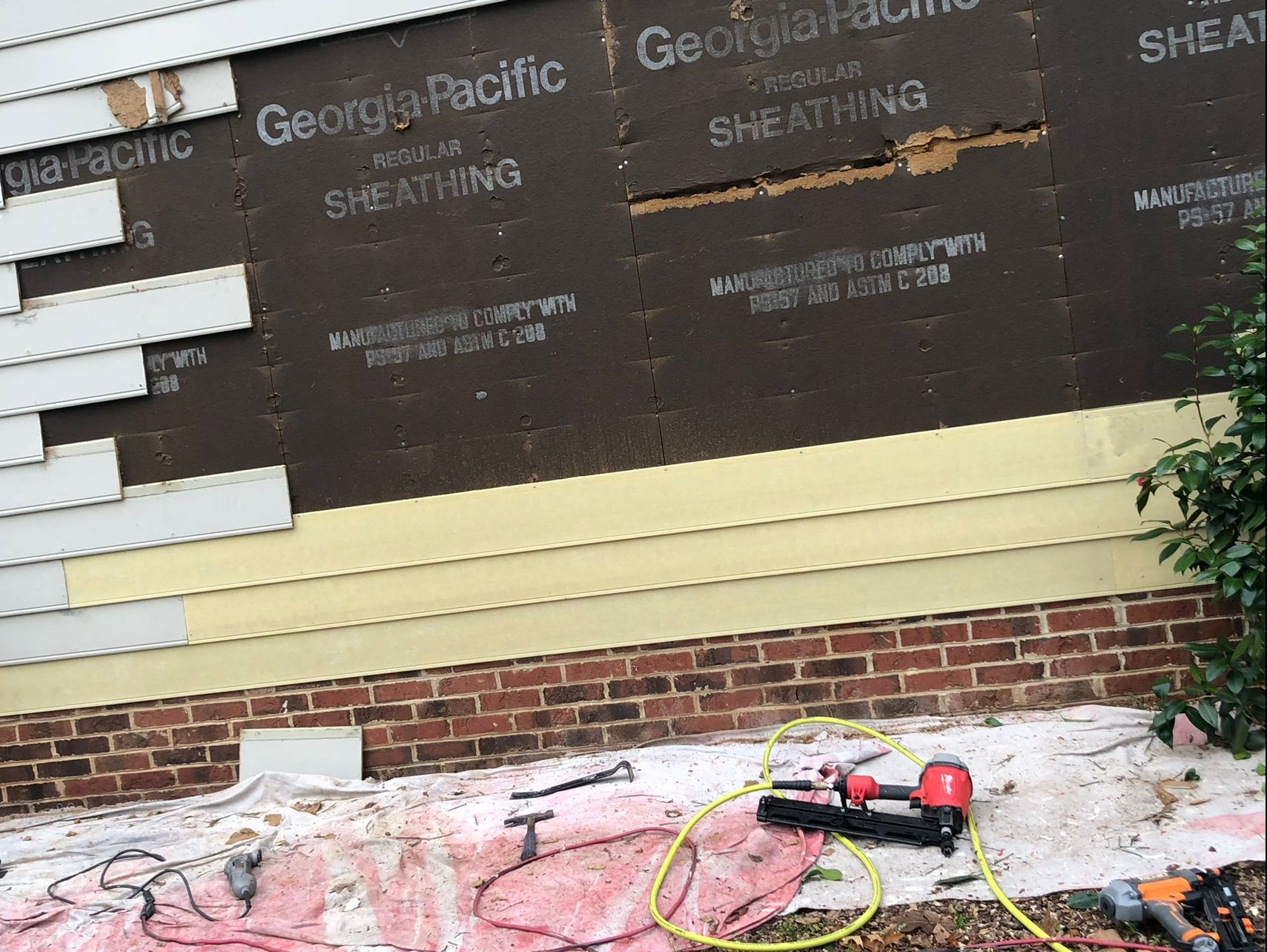 House siding partially replaced, showing new sheathing, siding, brick, and nail gun.