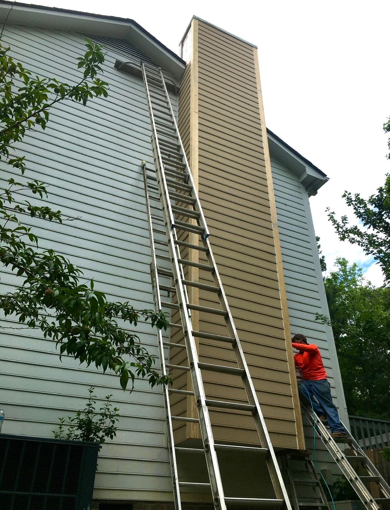 A person on a ladder working on a tall chimney attached to a light blue house.