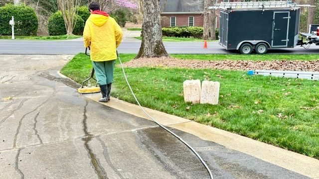 Person in yellow rain gear power washing a driveway. A trailer and ladder are also present.
