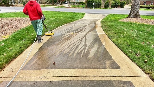 Person in red sweatshirt power washing a concrete driveway, leaving lighter streaks.