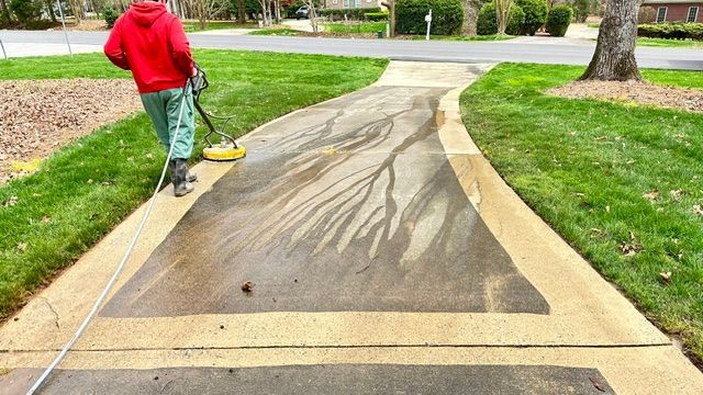 Person power washing a driveway, removing dirt and revealing clean concrete.