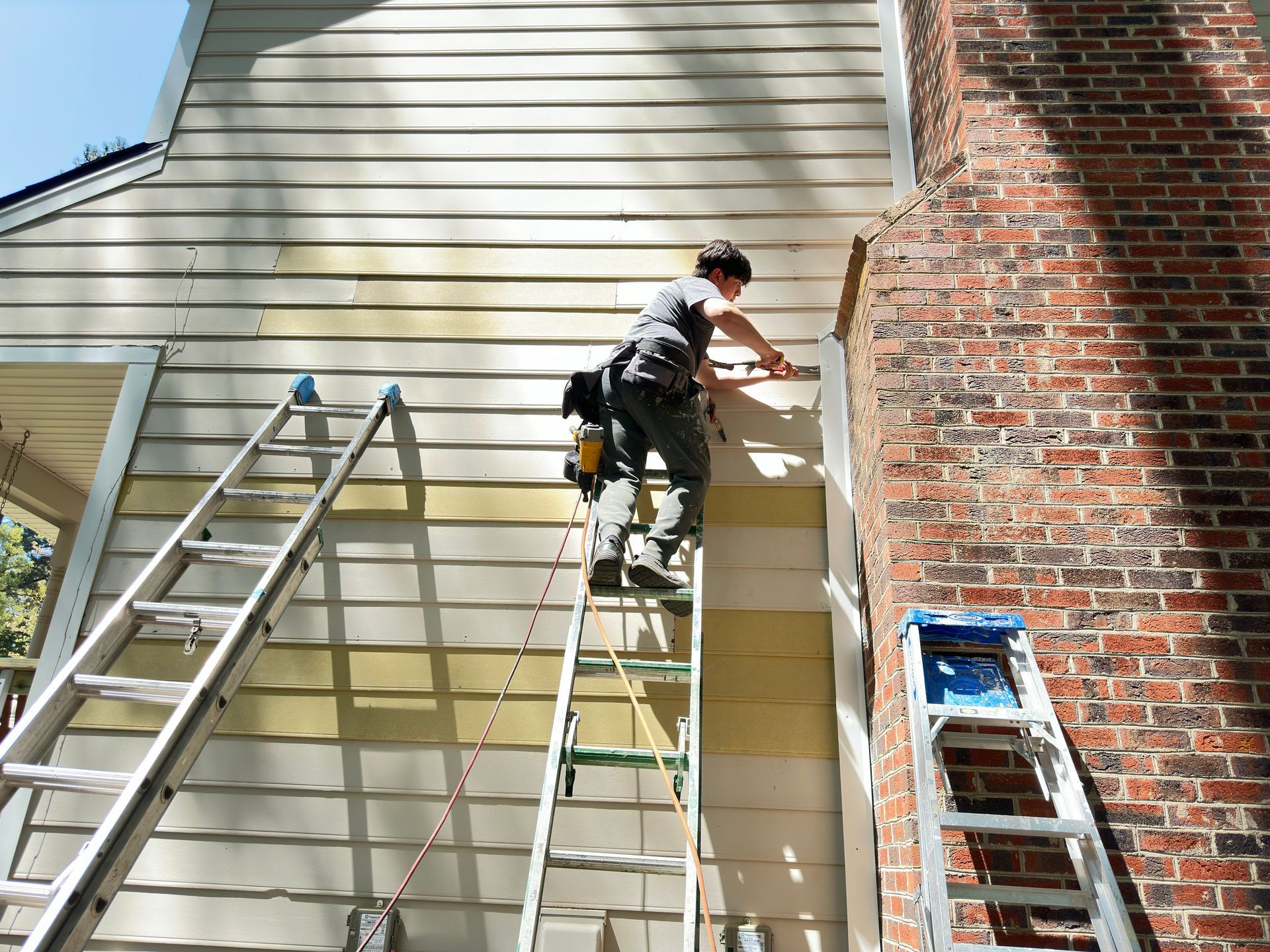 Person on ladder repairing siding near a brick chimney. Two other ladders are present.