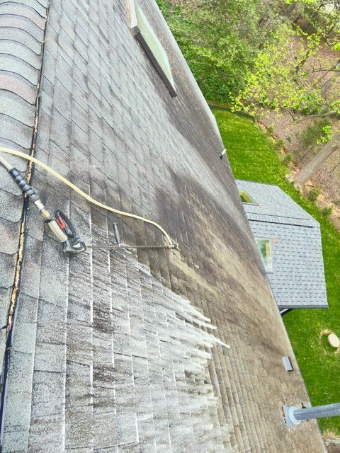 A person cleaning a dark roof with a pressure washer. A section of the roof is clean, revealing its original color.
