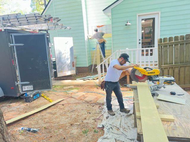 Construction workers using power saw near a house, with another worker on a ladder.