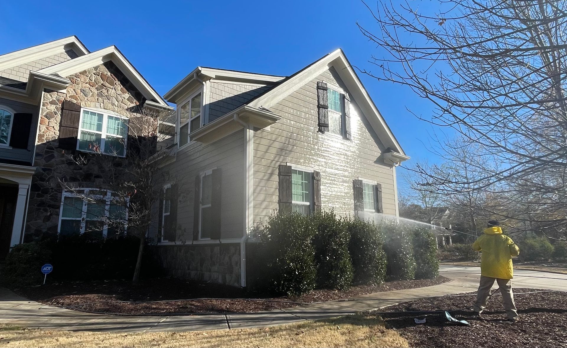 Person in yellow jacket pressure washing a brick house on a sunny day.