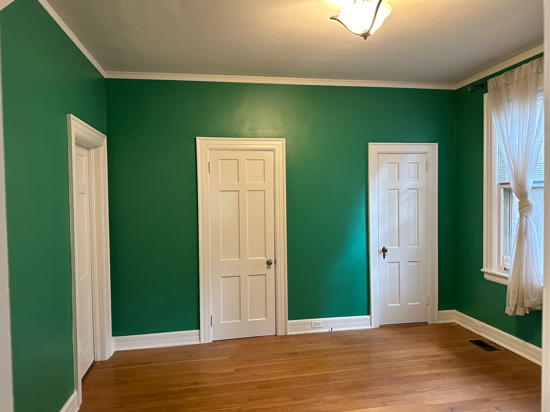 Empty room with green walls, white doors, trim, and light fixture, wood floor, and window with curtains.