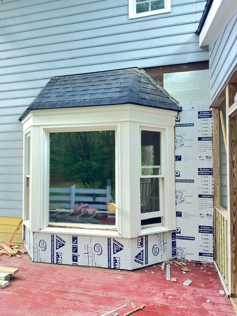 Bay window with white trim and dark roof under construction against gray house siding.