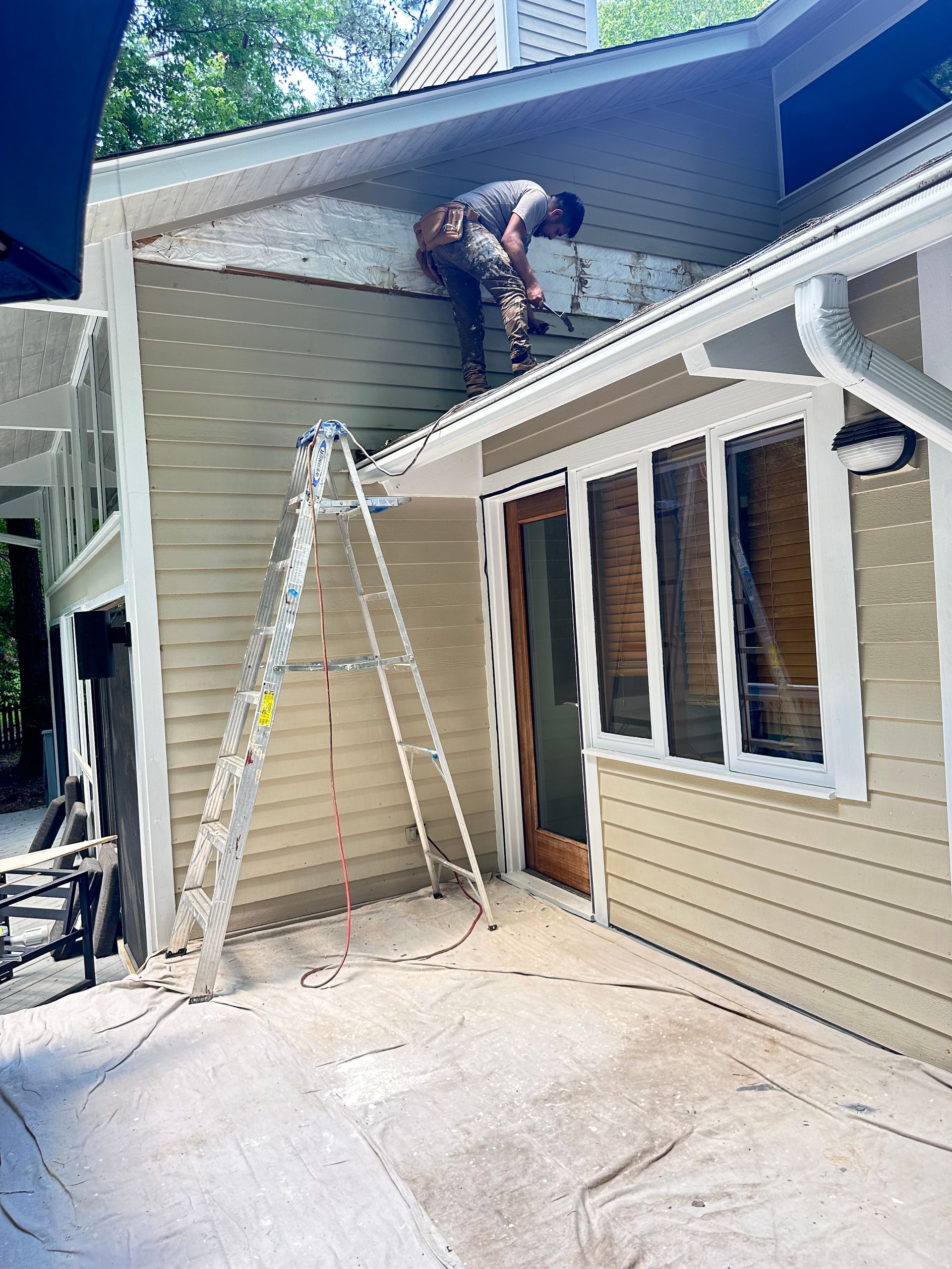 Man on ladder repairing siding on a house. Beige siding, wooden door, gray concrete patio.