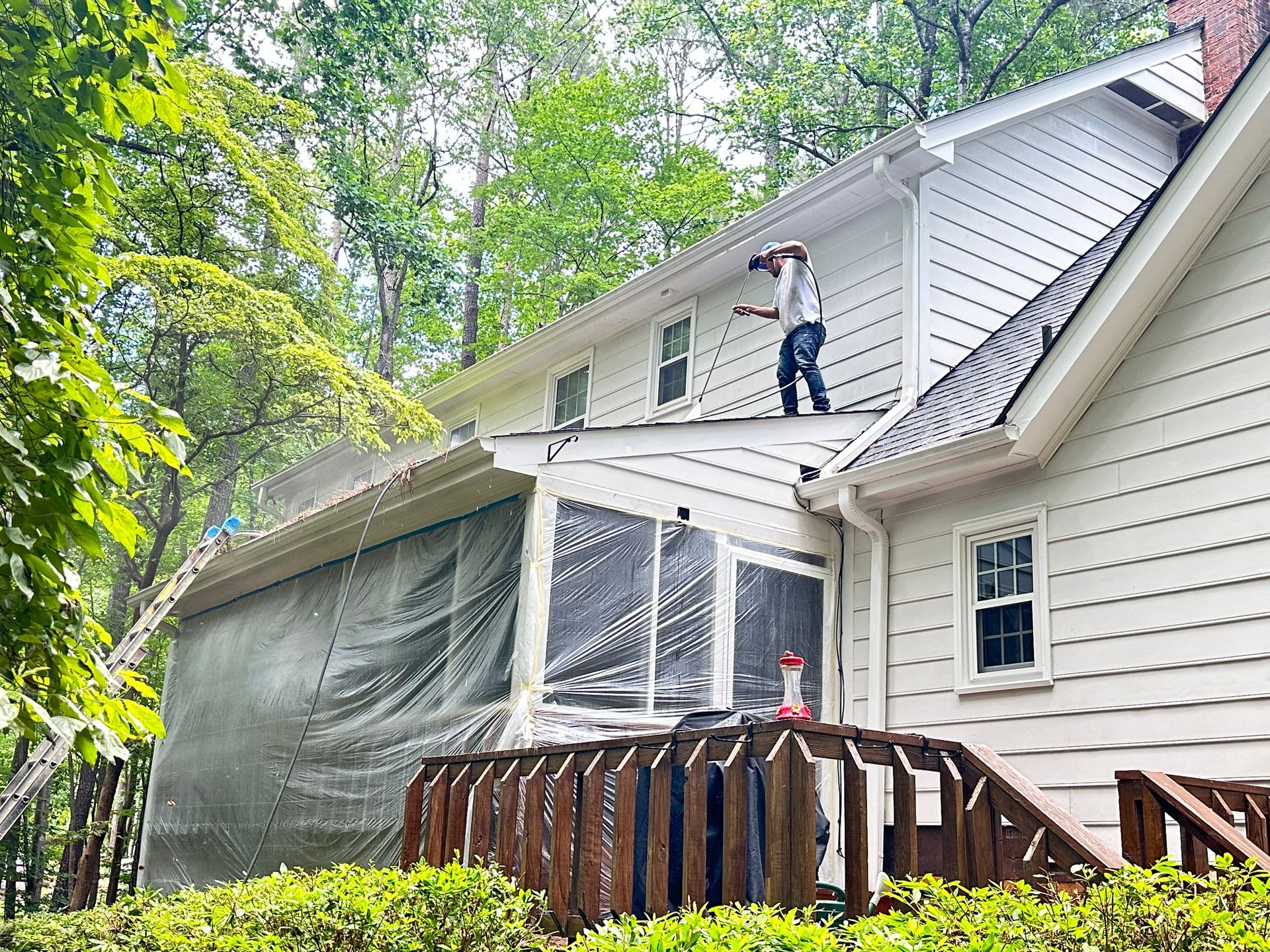 Person power washing a white house's siding, with a deck and enclosed porch visible. Trees in the background.