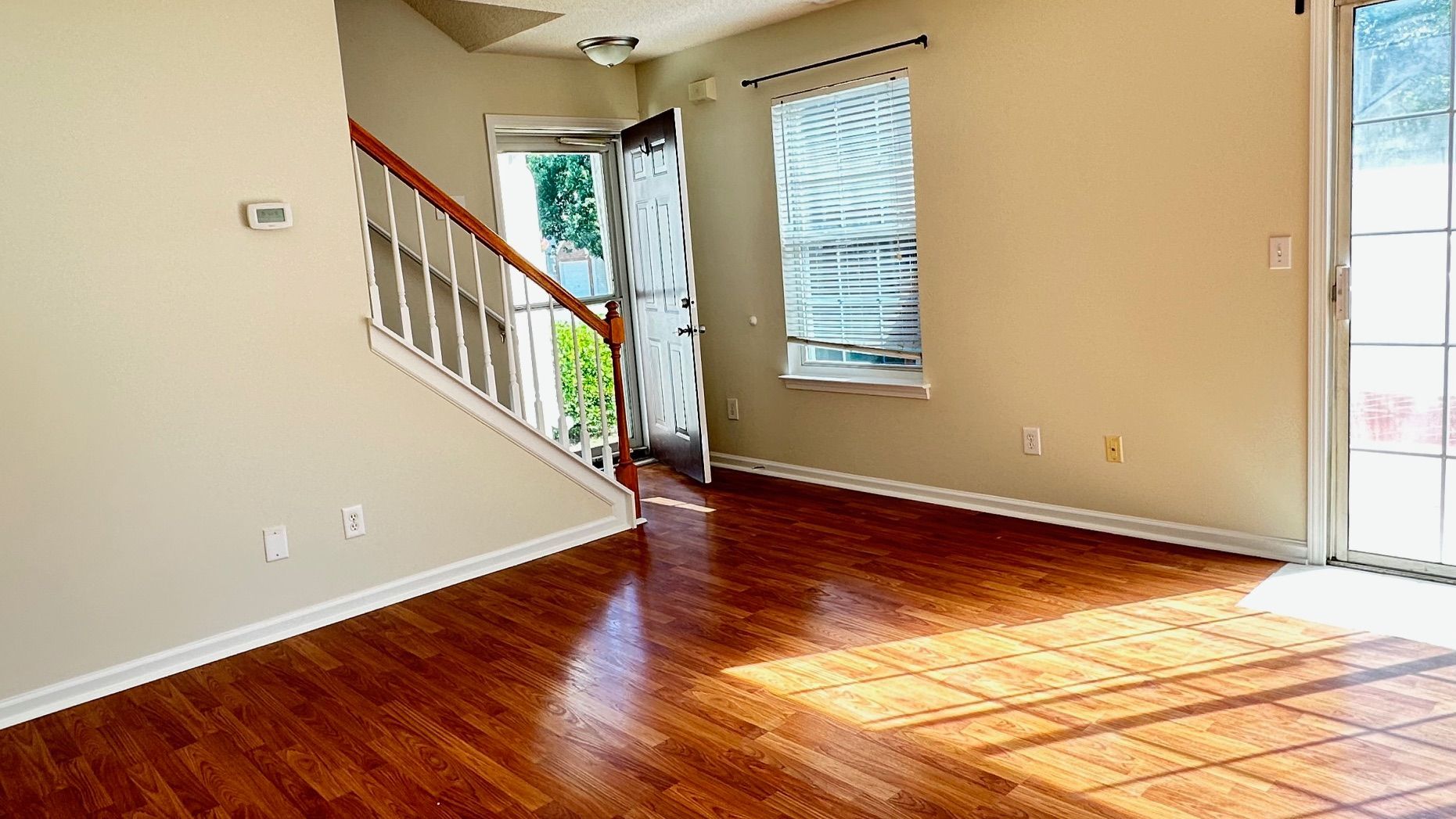 Empty living room with hardwood floors, staircase, and open doorway. Sunlight streams in from a