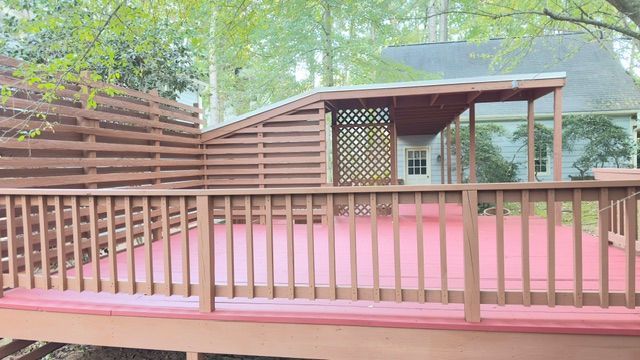 Wooden deck with red painted floor, brown railing, and latticework canopy. Trees and house in the background.