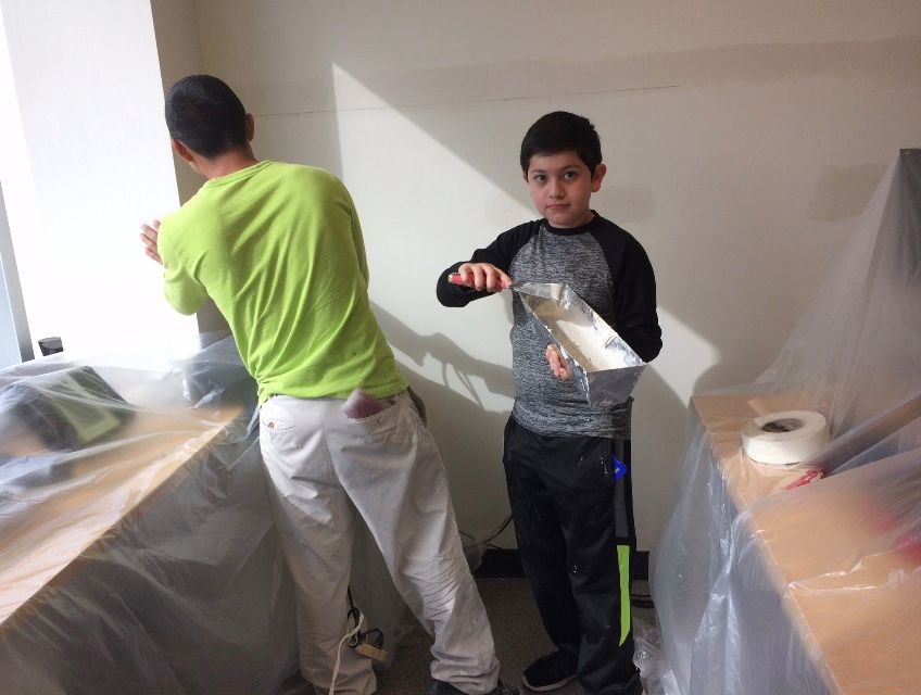 Two people applying drywall mud. One is on the wall, the other holding a tray. Room is under construction.