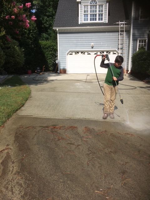 Person power washing a driveway in front of a blue house with a white garage door.