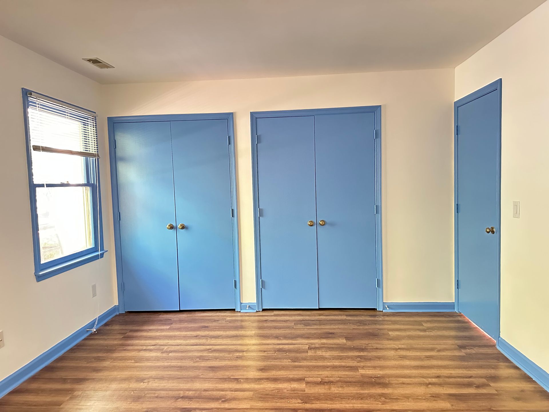 Empty room with three blue doors, a window, and wood flooring. Walls are white and beige.