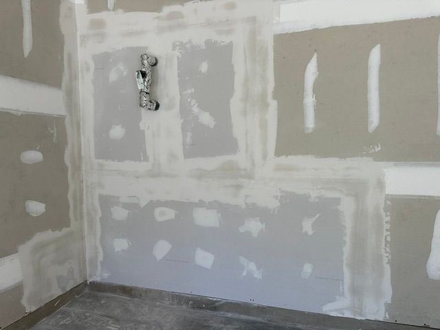 Drywall installation in progress, with taped seams and electrical box visible. Gray walls and flooring.