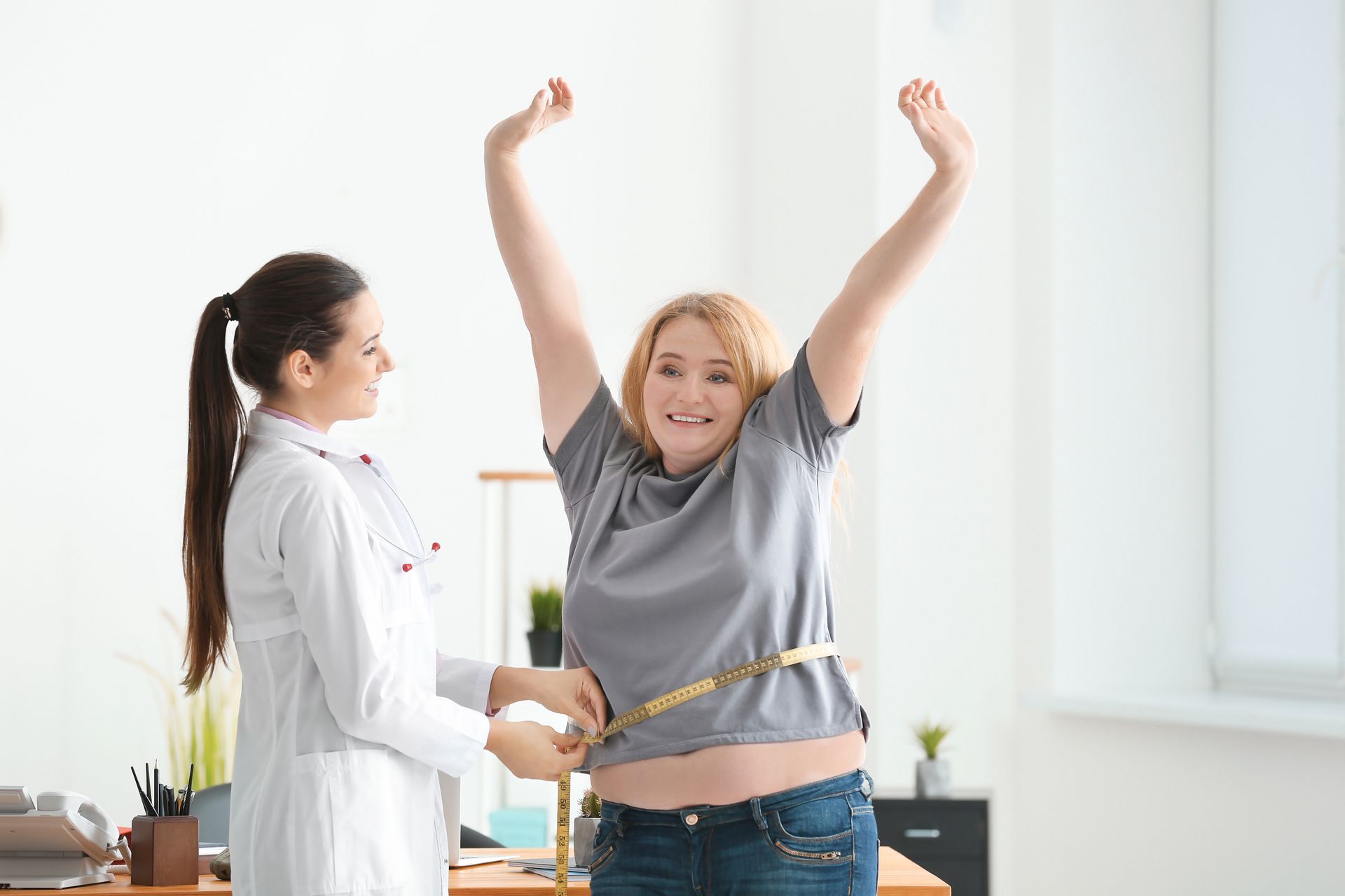 Person raising arms while another measures waist with tape in a bright room.