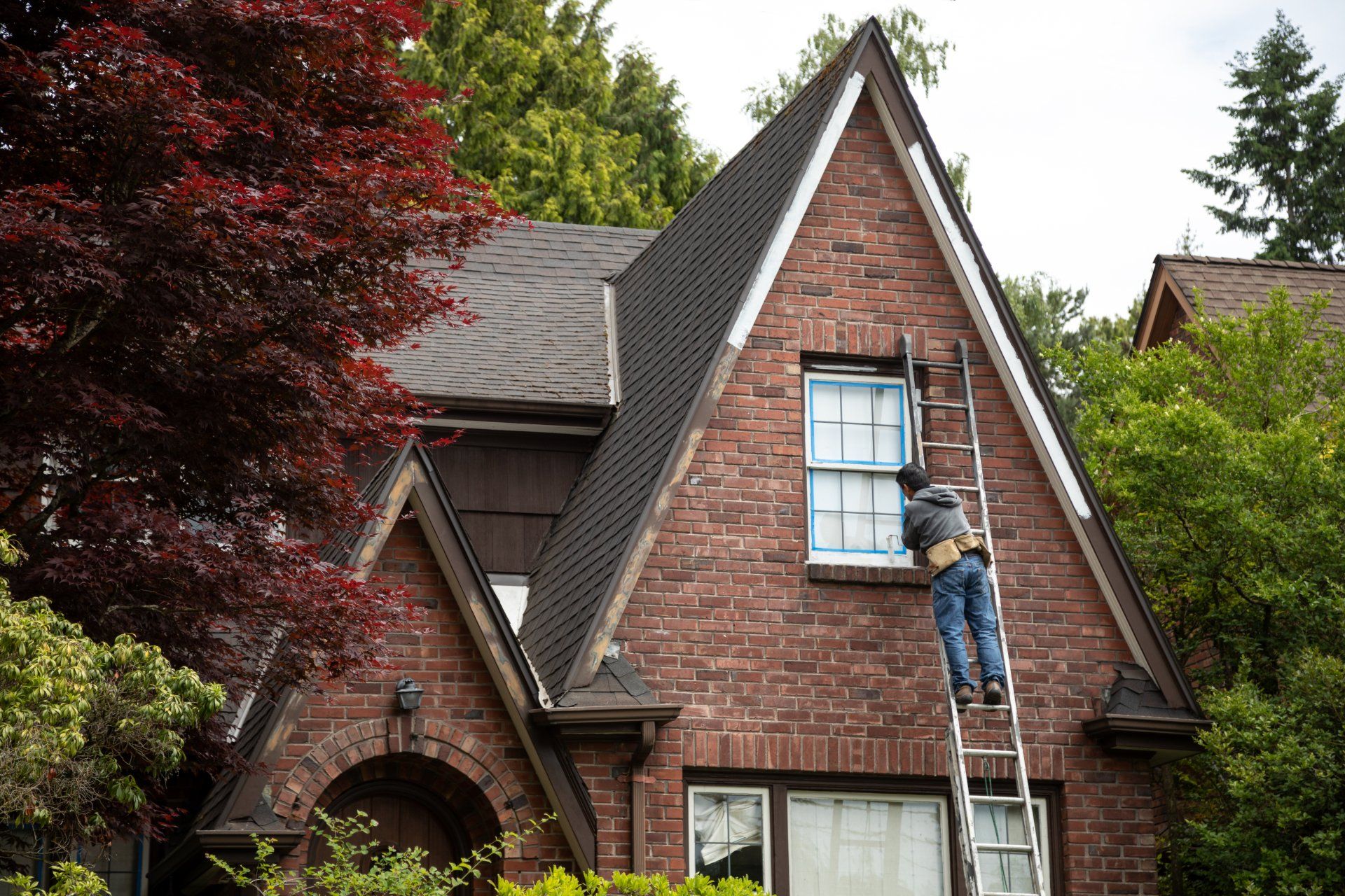 A man on a ladder is painting a window on a brick house.