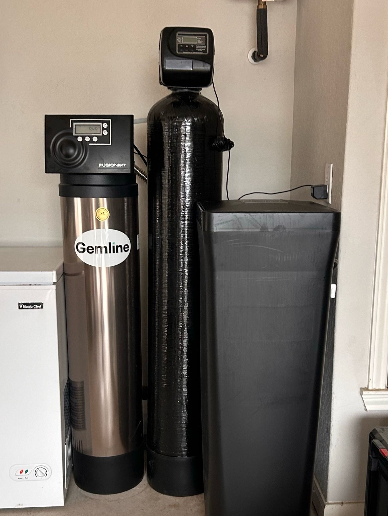 Water filtration system components in a utility room. Three tanks of varying sizes and a white box are against a wall.