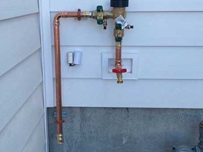 Copper piping and a backflow preventer assembly mounted on a white exterior wall with a red-handled valve.
