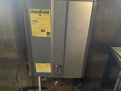 A grey tankless water heater mounted on a wall with two yellow EnergyGuide labels and a digital display showing 120.
