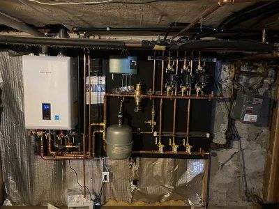 Wall-mounted boiler and heating manifold system with copper piping and grey expansion tanks in a basement.