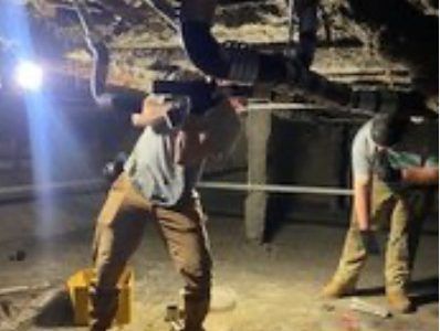 Two people in work clothes repair pipes in a dimly lit crawl space under a house.