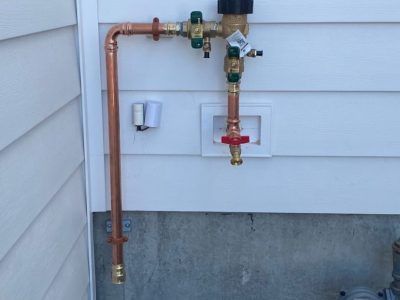Copper piping on a white vinyl-sided wall, featuring a vacuum breaker valve and a water spigot in a recessed box.