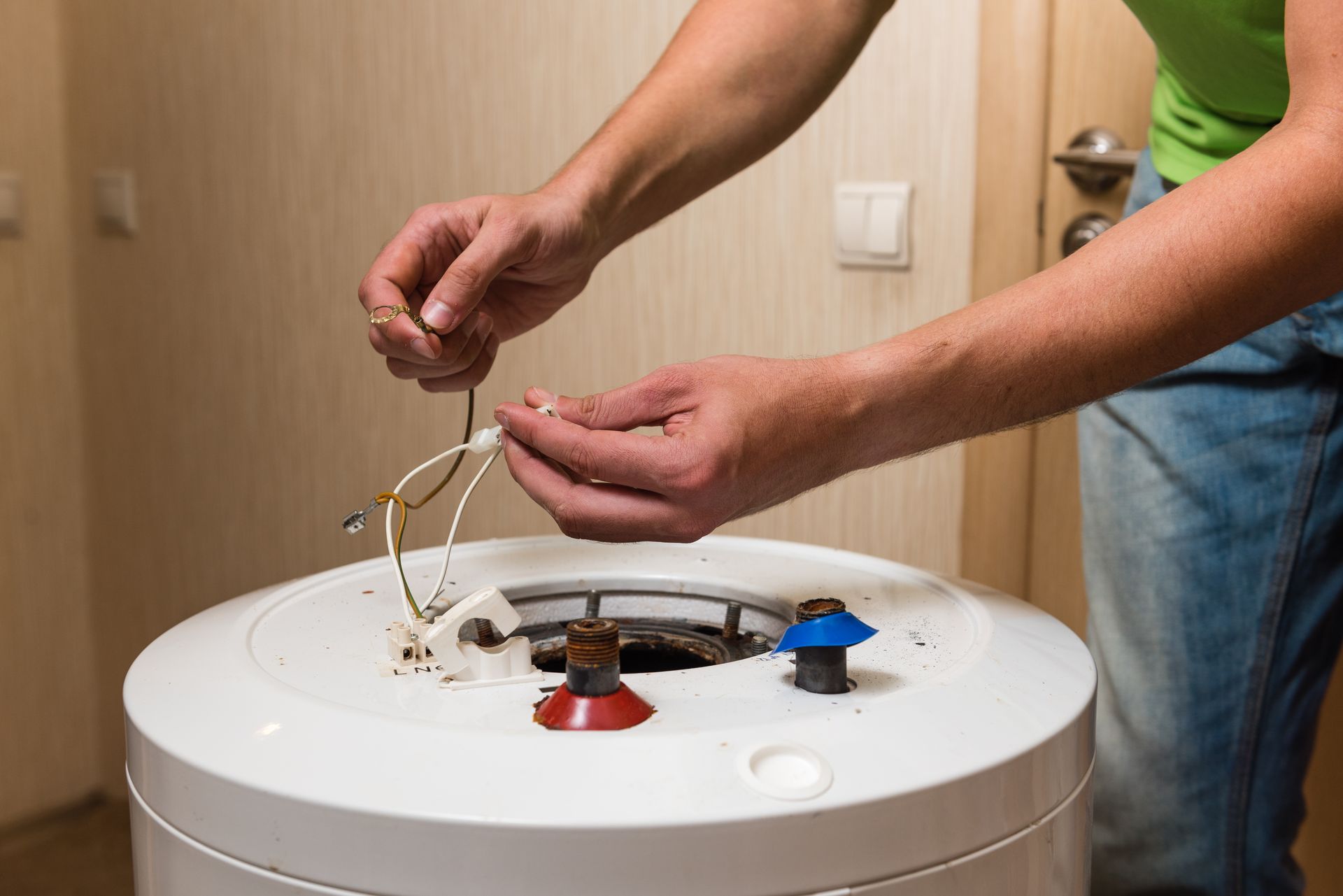 A technician works on the open top of a white electric water heater, handling internal wiring.