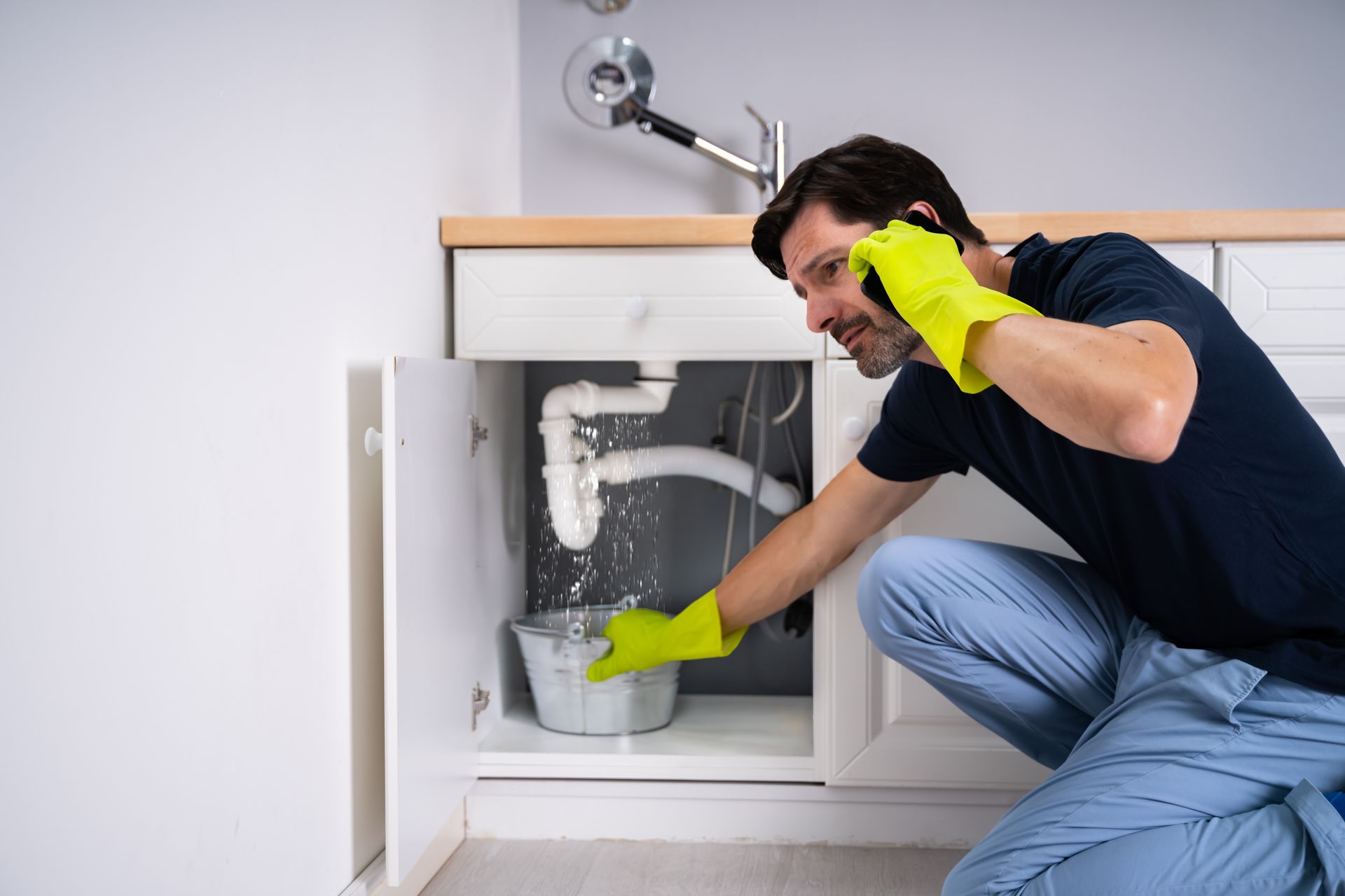 A person wearing yellow rubber gloves talks on a phone while holding a bucket under a leaking sink pipe.
