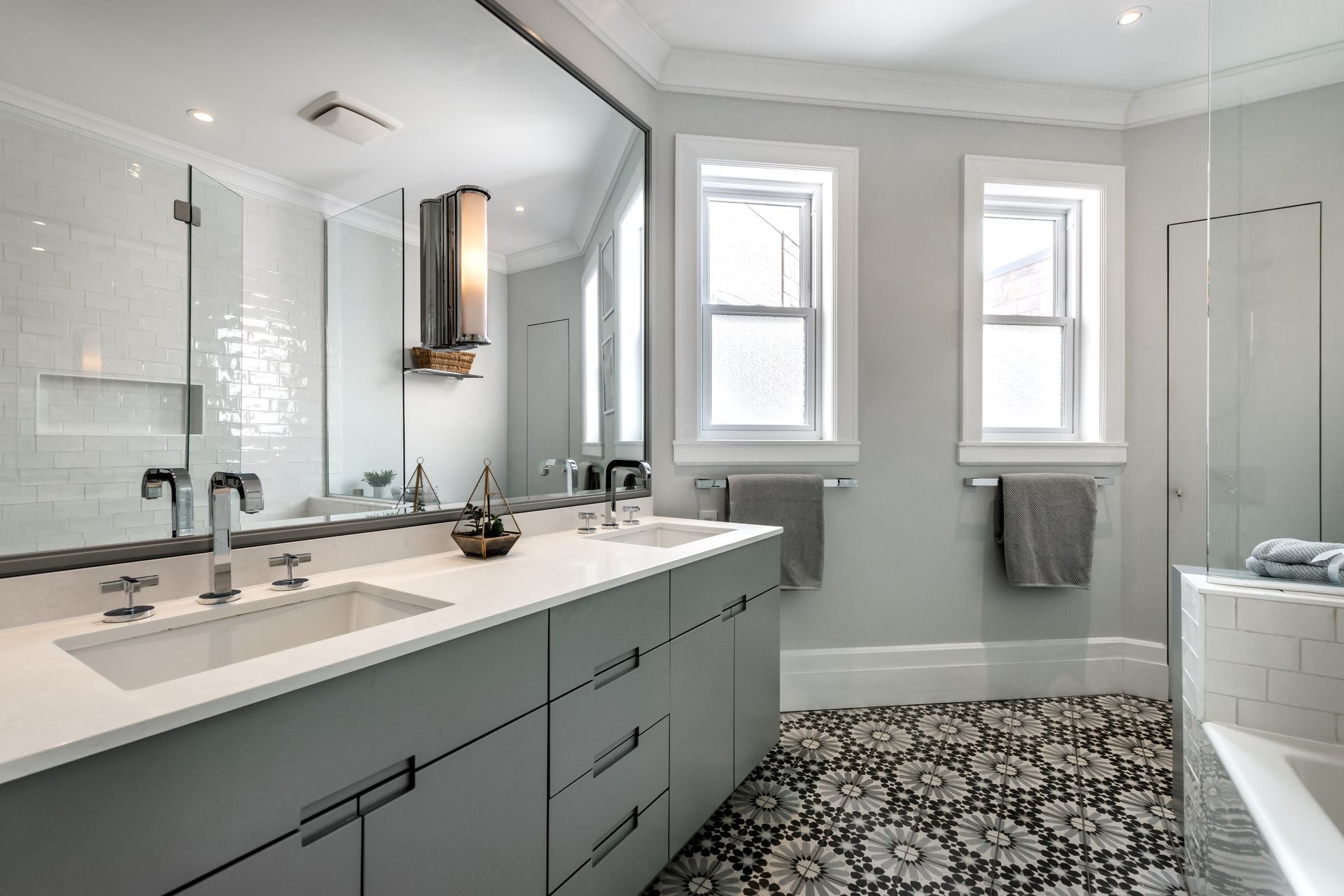 A modern bathroom with a long grey double vanity, white countertops, two windows, and patterned black and white flooring.