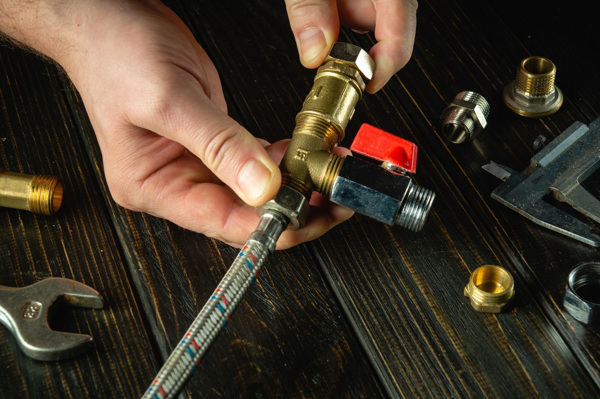 Hands assemble brass plumbing fittings and a flexible hose on a dark wood surface with various tools nearby.