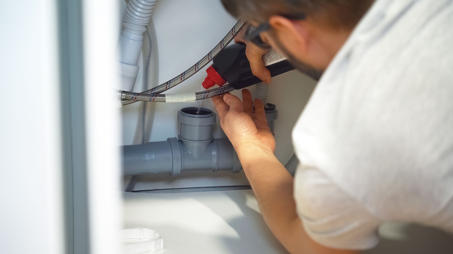 A person in a white shirt installs gray PVC plumbing pipes under a sink.