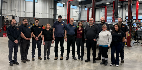 Group of people posing in an auto repair shop. They wear work uniforms and stand near car lifts.