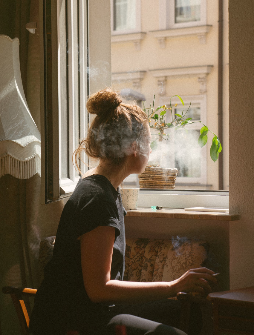 Woman exhales smoke by a window, looking outside. Soft sunlight. Building in the background.