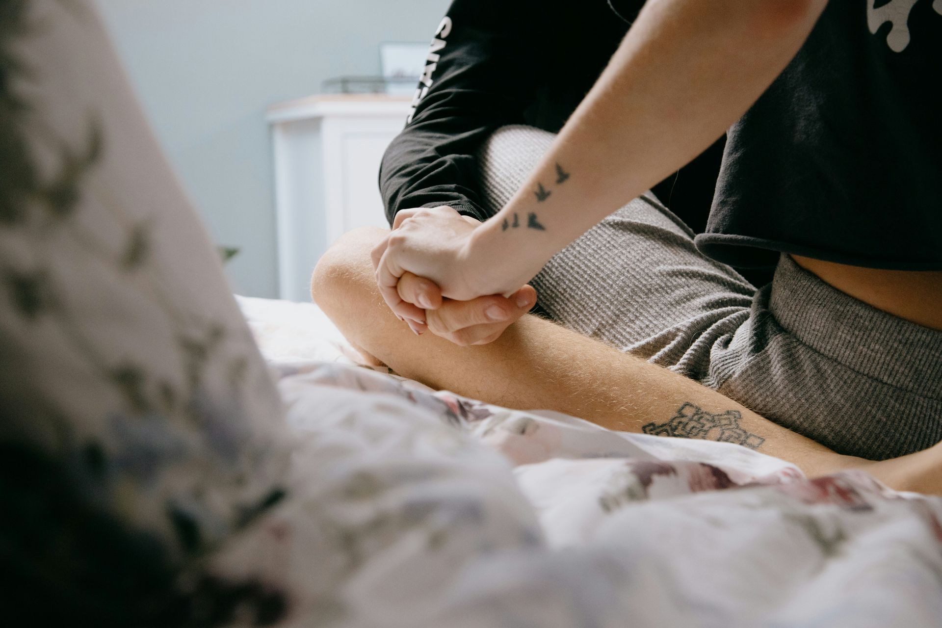 Two people holding hands on a bed, tattoos visible, soft lighting.