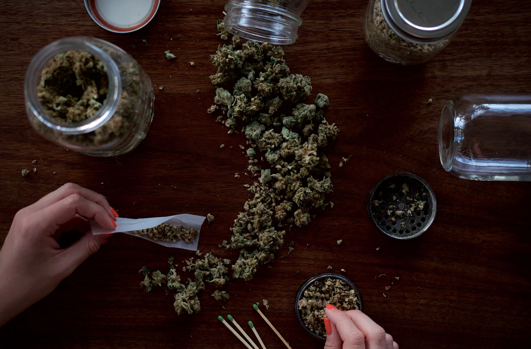 Hands rolling a joint with scattered cannabis buds and jars on a wooden surface.