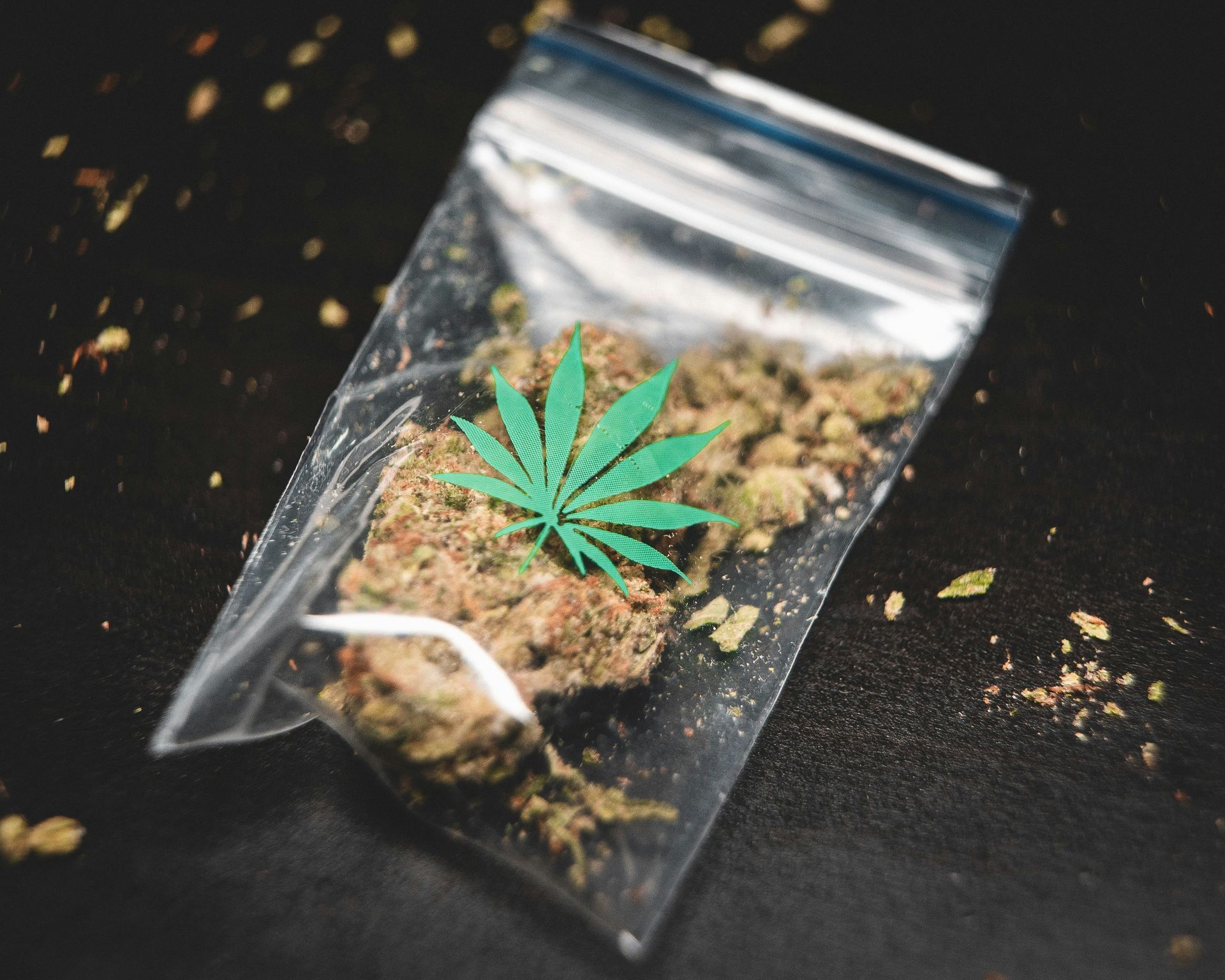 Marijuana buds in a clear plastic bag with a green cannabis leaf on a dark surface.