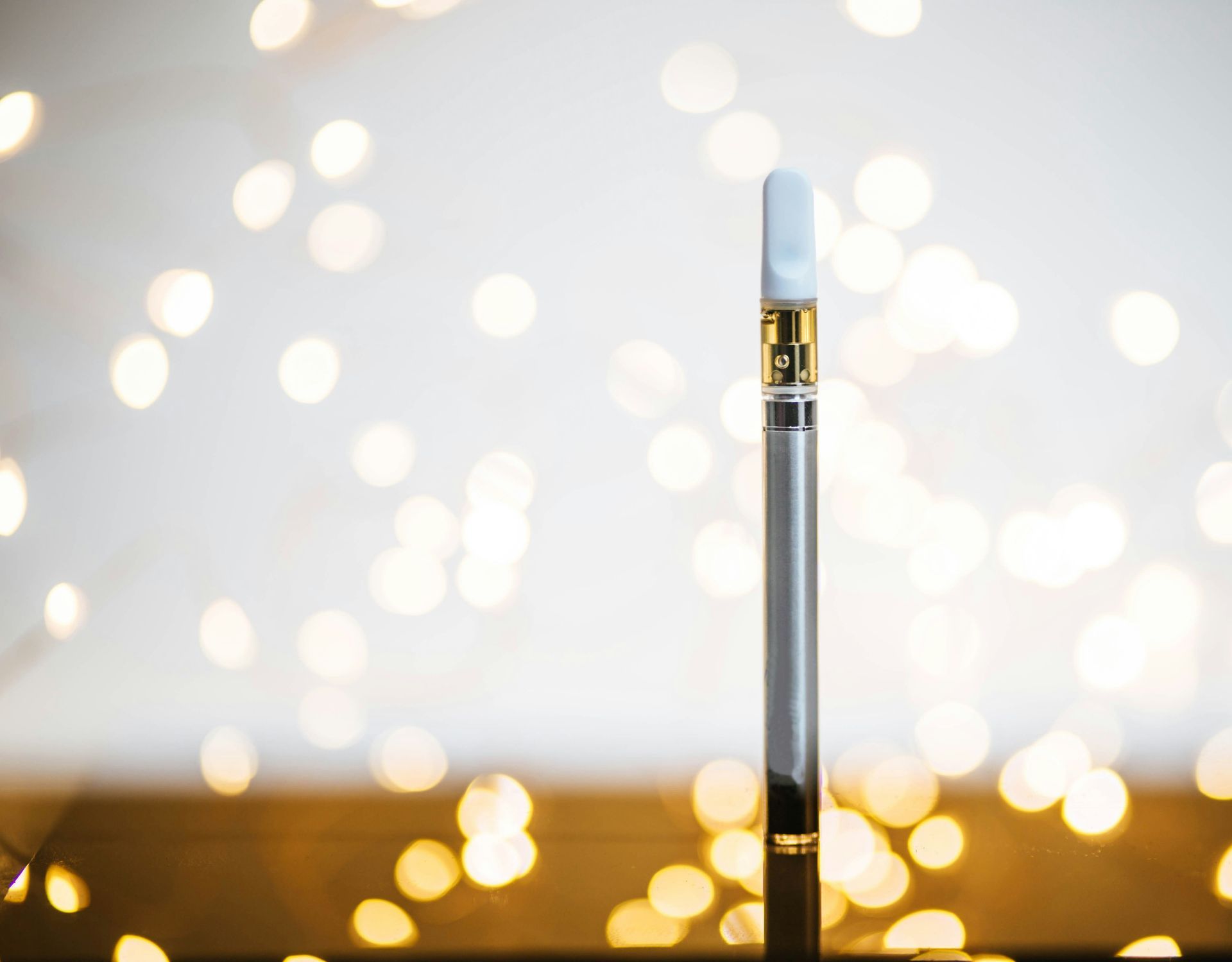 Vape pen, silver body, white mouthpiece, against a blurred background of golden bokeh lights.