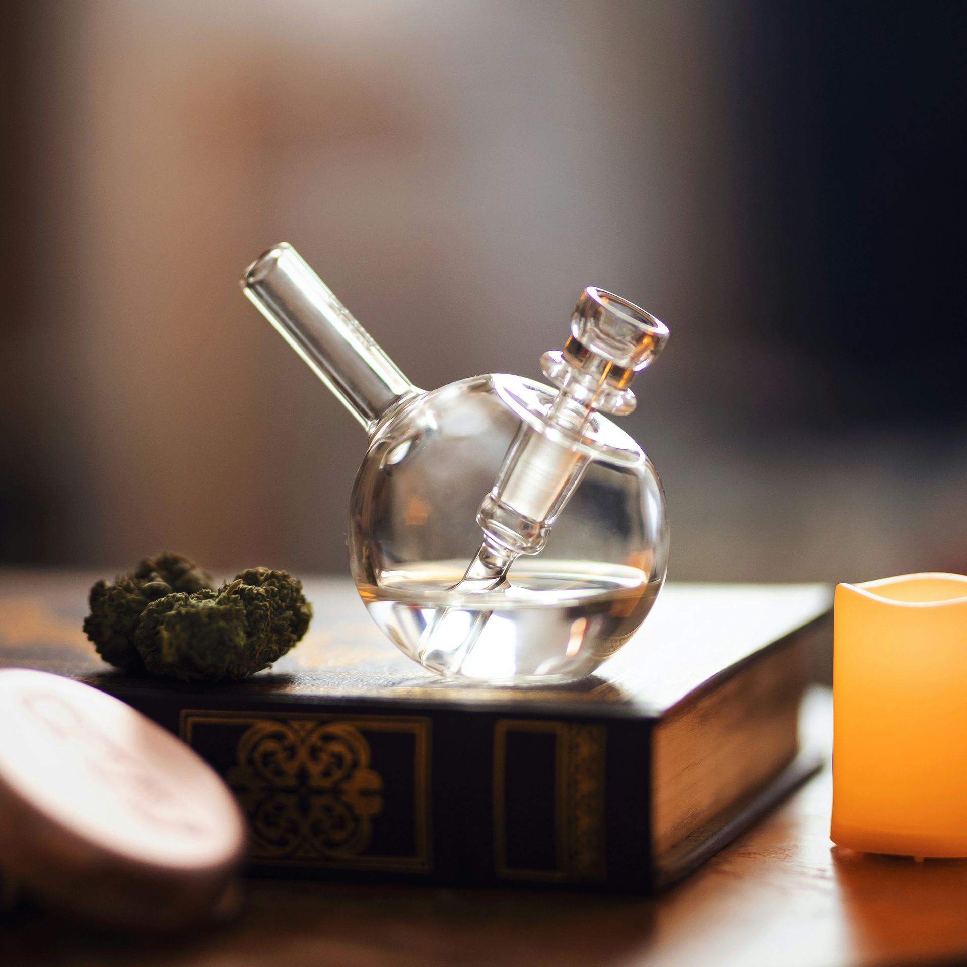 Small glass bong with water, cannabis bud, book, and candle.