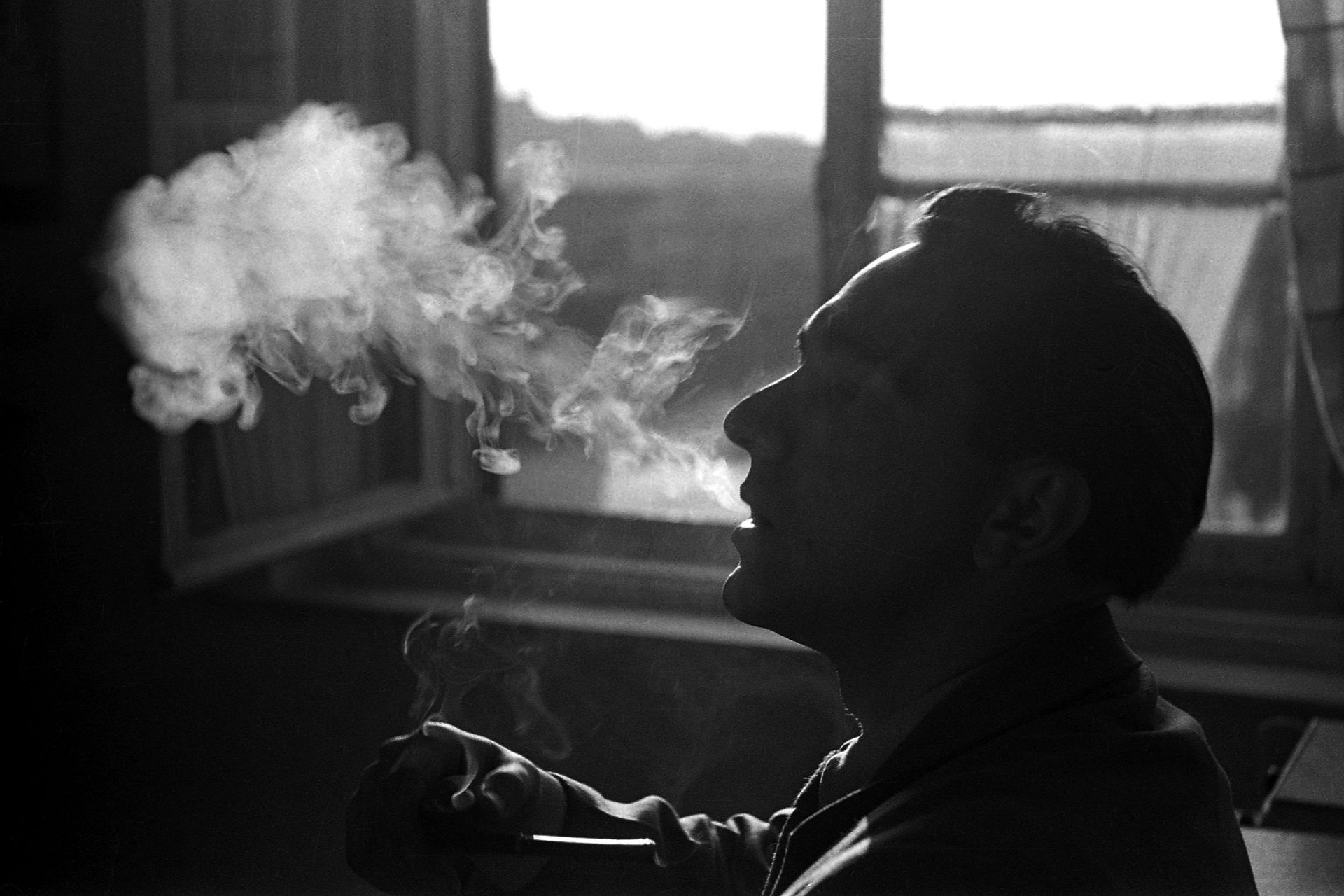 Man exhales a plume of smoke from his mouth in a room near an open window.