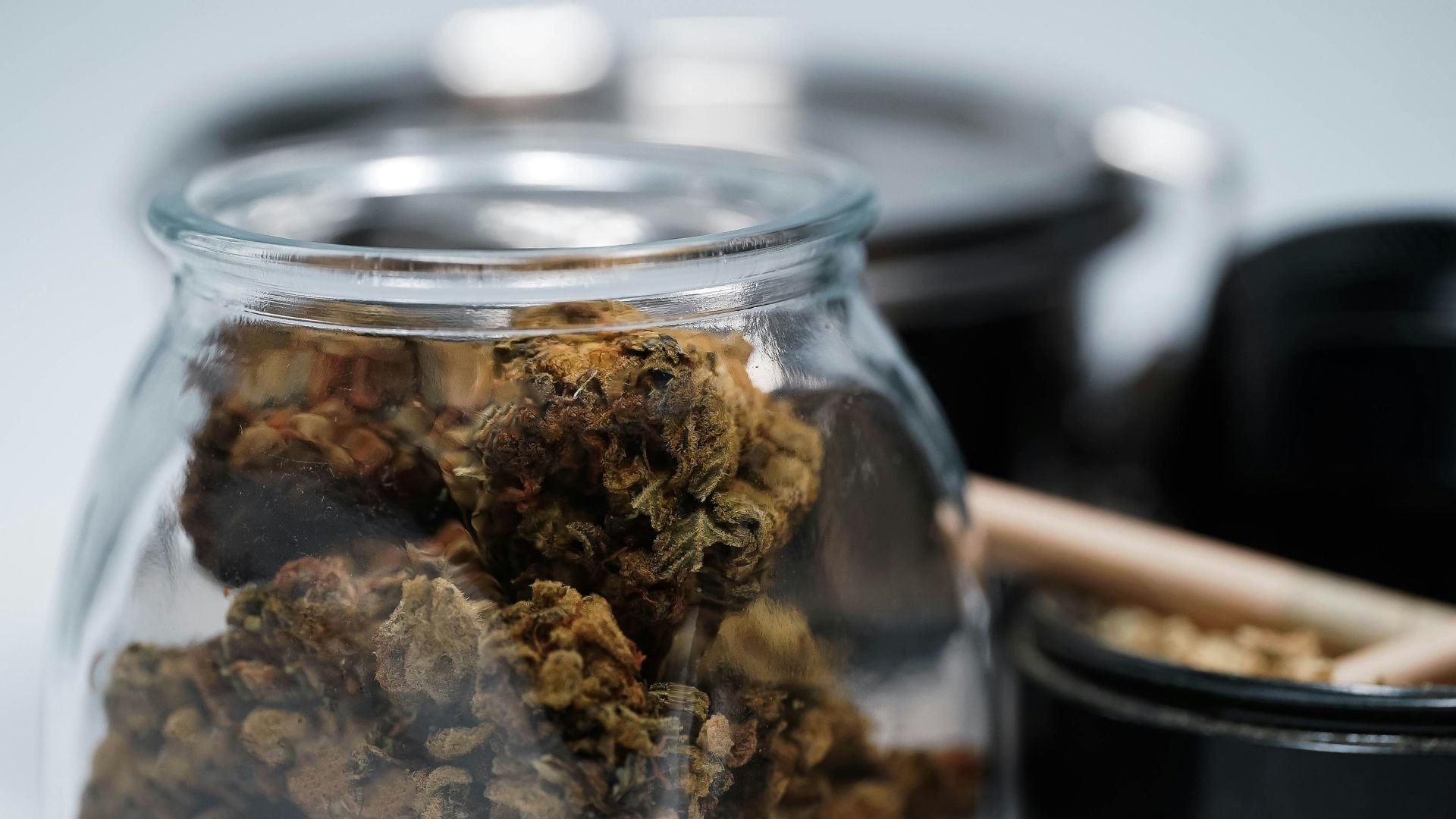 A jar filled with cannabis buds, in focus. Additional items include a black bowl and other dark items.