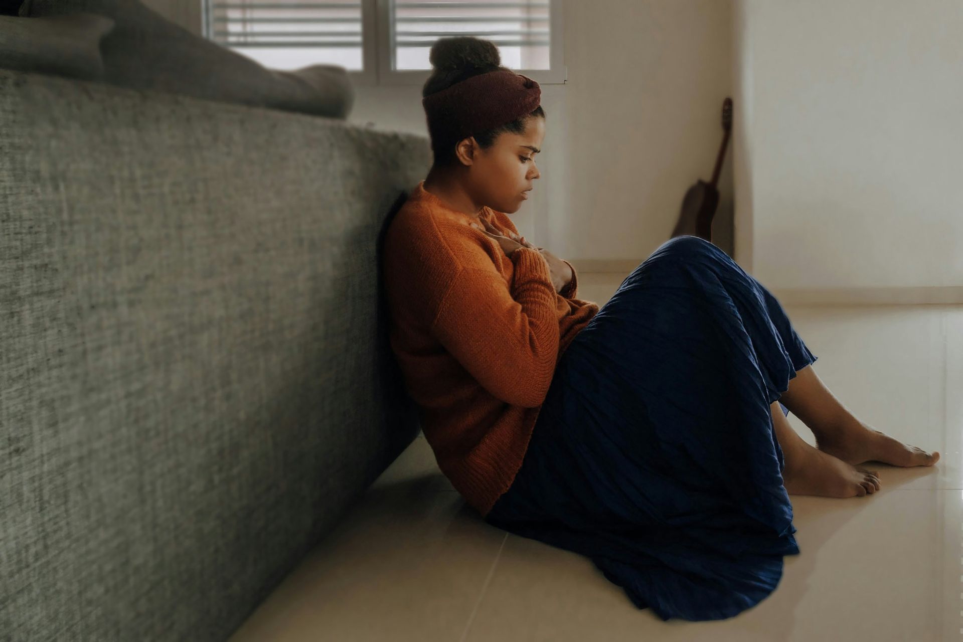 Woman sits against a wall, looking down, hands clasped, wearing orange sweater and blue skirt.